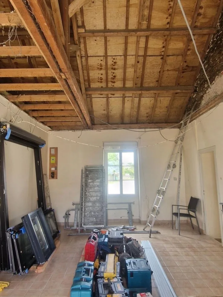 Pièce en rénovation au plafond déposé, solives en bois apparentes, avec échelle, échafaudage, caisses à outils et fenêtres neuves en attente de pose