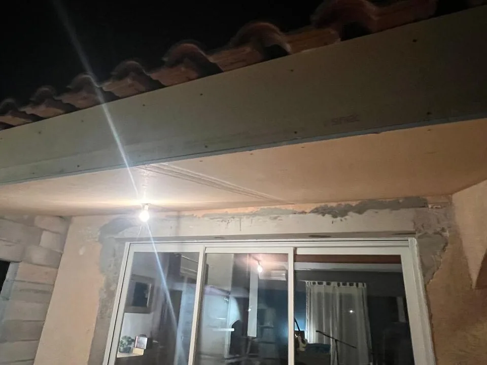 Sous-face de toiture habillée en plaques au-dessus d'une baie vitrée coulissante, photographiée de nuit sous un éclairage de chantier