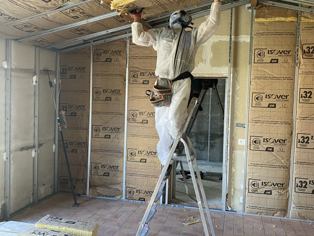 Artisan en combinaison et masque de protection, sur un escabeau, posant l'isolant du plafond entre les rails métalliques, murs déjà garnis de panneaux isolants